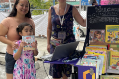 Two women and a smiling child pose in front of a pop-up library at an outdoor event; the young girl is holding a book she selected.
