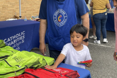 A young boy standing with a volunteer while selecting a backpack at an outdoor event.