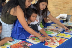 Three children browsing through books on a table at an outdoor event.