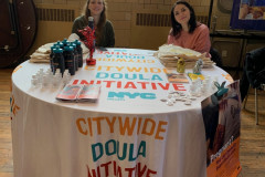 Two smiling women seated at a round table cover in a table cloth from the NYC Doula Program.