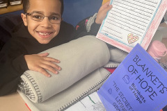 Smiling boy holding a gray blanket and a handwritten note next to a Blanket of Hope flyer.