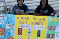 Two women sitting at a table with a sign about healthy alternatives for beverages and snacks.