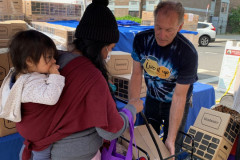 A member of Catholic Charities Brooklyn and Queens Community Wellness team stands under a blue tent and distributes a box of fresh produce to a mom carrying a child on her back.