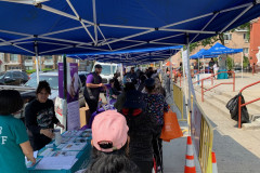 A group of people standing under a blue tent as they receive information and resources and a box pf fresh produce from Catholic Charities Brooklyn and Queens.
