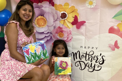 A woman and young girl hold floral paintings on canvas in front of a decorative Mother's Day backdrop.