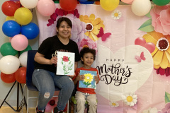 A woman and young girl hold floral paintings on canvas in front of a decorative Mother's Day backdrop surrounded by colorful balloons.