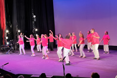A group of women  dressed in pink shirts and white pants dancing on stage in front of an audience.